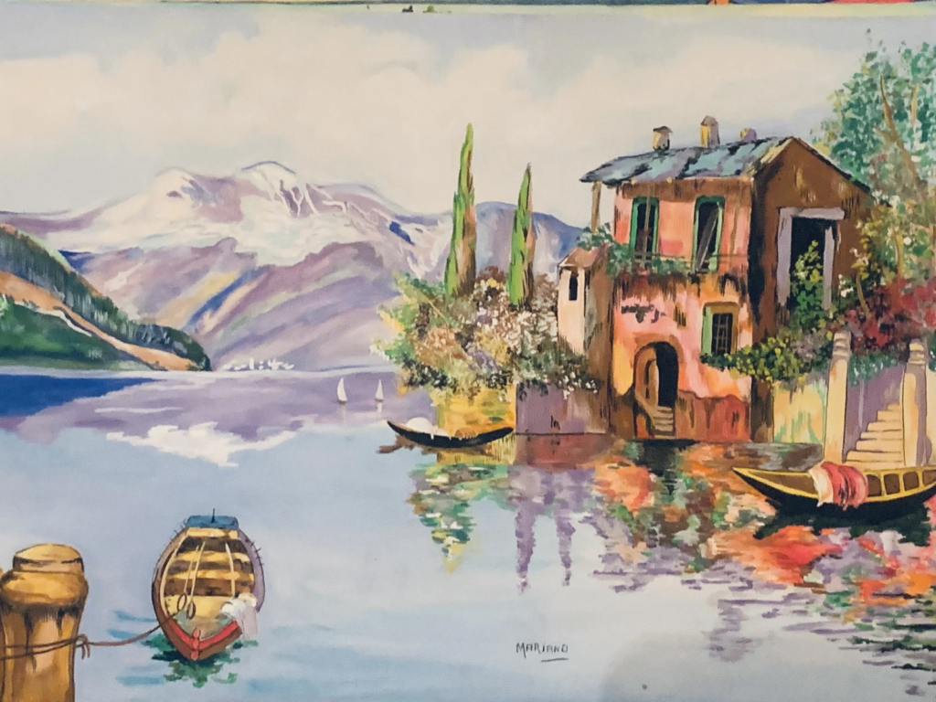 A lakeside house, small boats and reflections, with pale mountains in the distance.