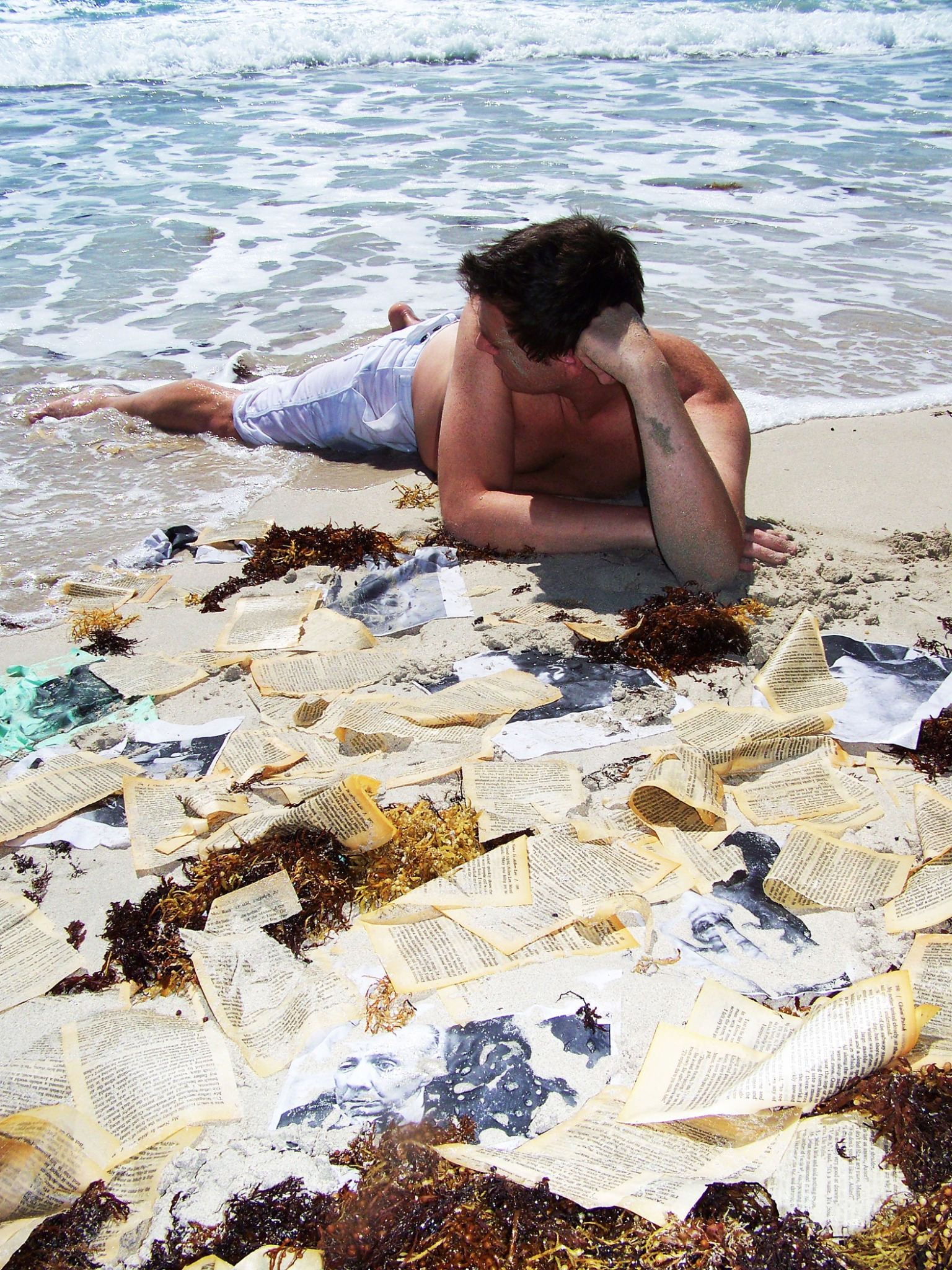 Picos Pelegri reclining on the beach beside scattered book pages and photographs.